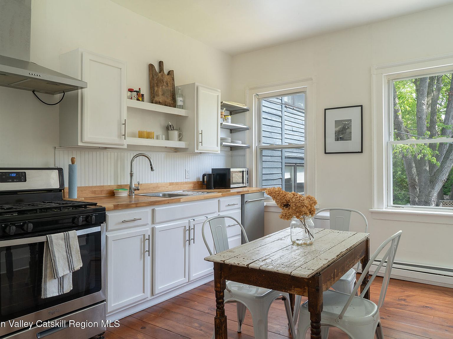 Renovated kitchen with wood-block counters, open shelving, white cabinetry, and original wide-plank pine flooring; light pours through tall single-pane windows over a rustic wood breakfast table