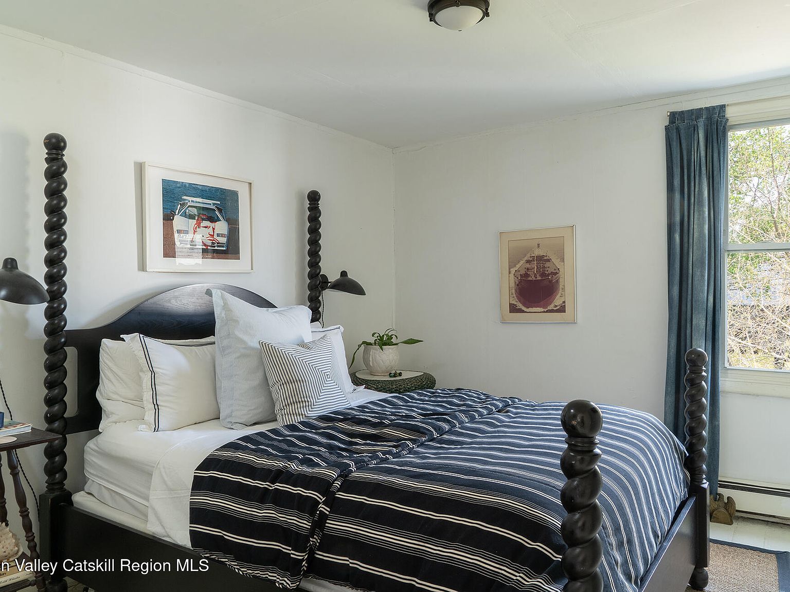 Guest bedroom with spool four-poster bed, navy ticking-stripe duvet, framed nautical art, and indigo drapes pulled back from a window looking onto leafy trees